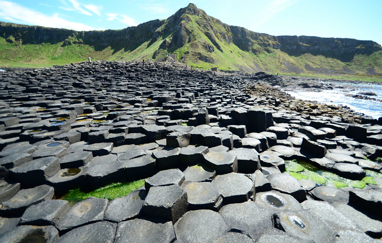 Giants Causeway with greenery and hills in the background against a bright blue sky with stones on the floor like a pathway