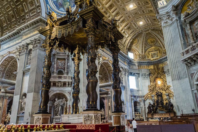 Een weergave van de binnenkant van de Sint-Pietersbasiliek naar boven gericht met de Sint-Pietersbasiliek (Italiaans: Baldacchino di San Pietro in focus met een gigantisch overweldigend gewelfd plafond op de achtergrond.