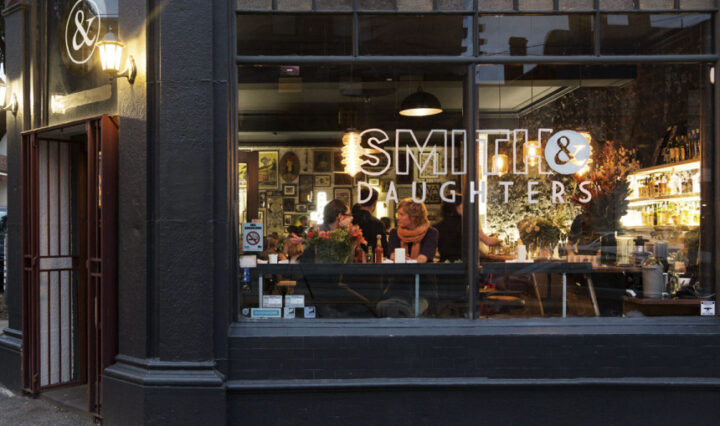 Голям прозорец с надпис „Smith & Daughters“ отпред и хора, които се хранят на маси в добре осветена стая