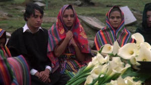 Two teenagers and a middle-aged woman sit in front of a flower-covered casket. 