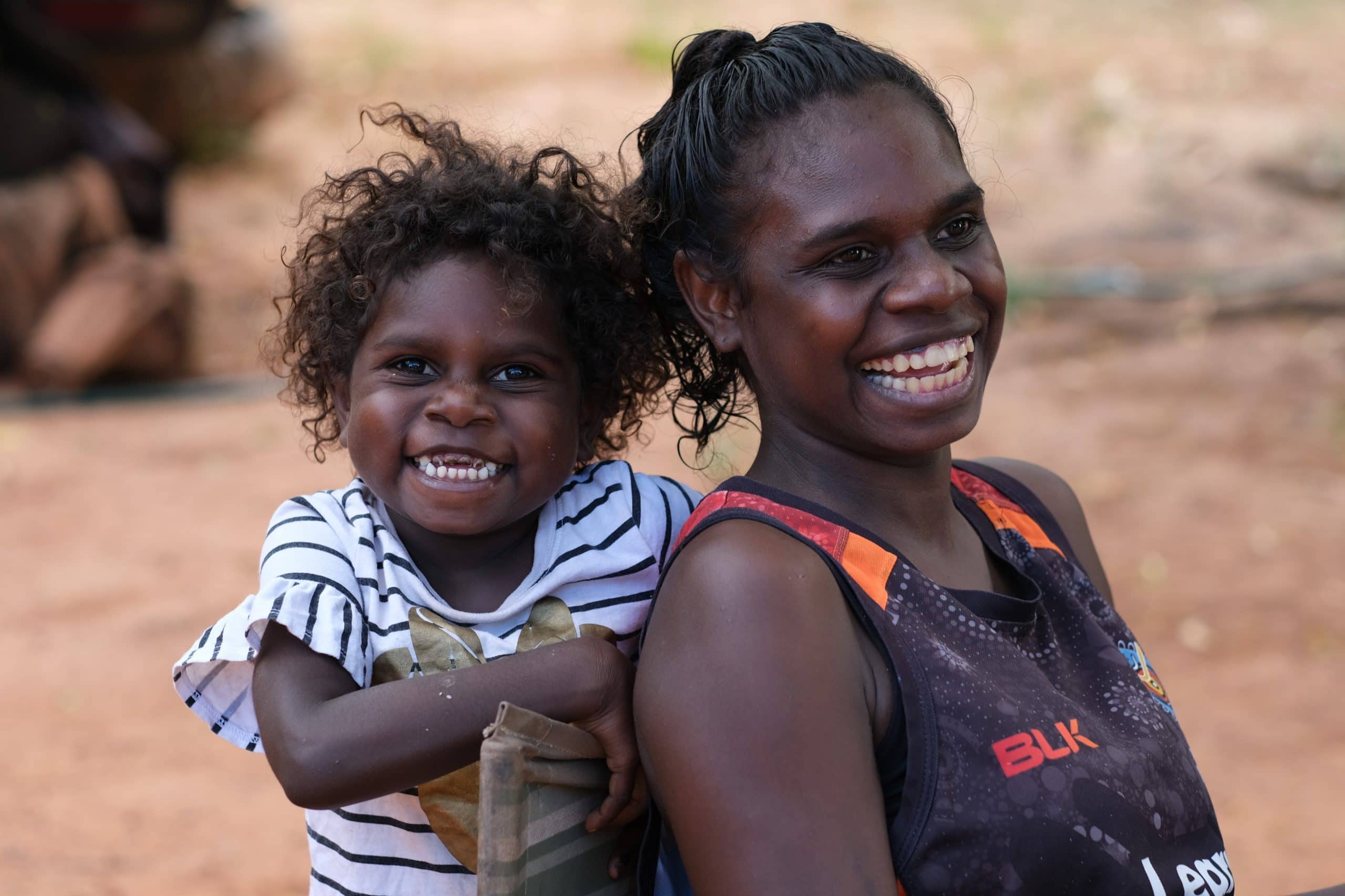 Изстрел отблизо на две млади аборигенски момичета, разположени рамо до рамо с усмивки на лицата им с оранжев пясъчен фон