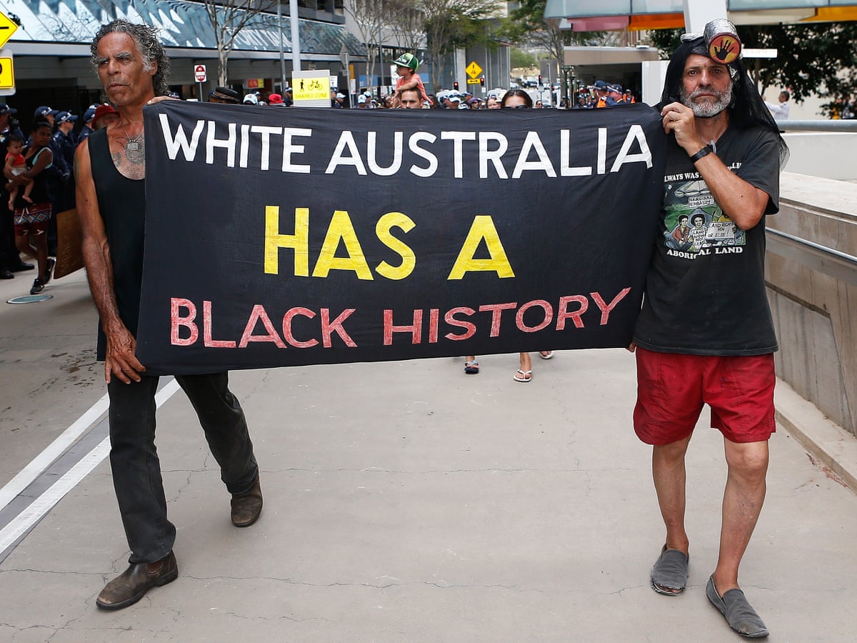Двама аборигени вървят към камерата, държейки черен плакат с текст „Бяла Австралия има черна история“, написан в бяло, жълто и червено