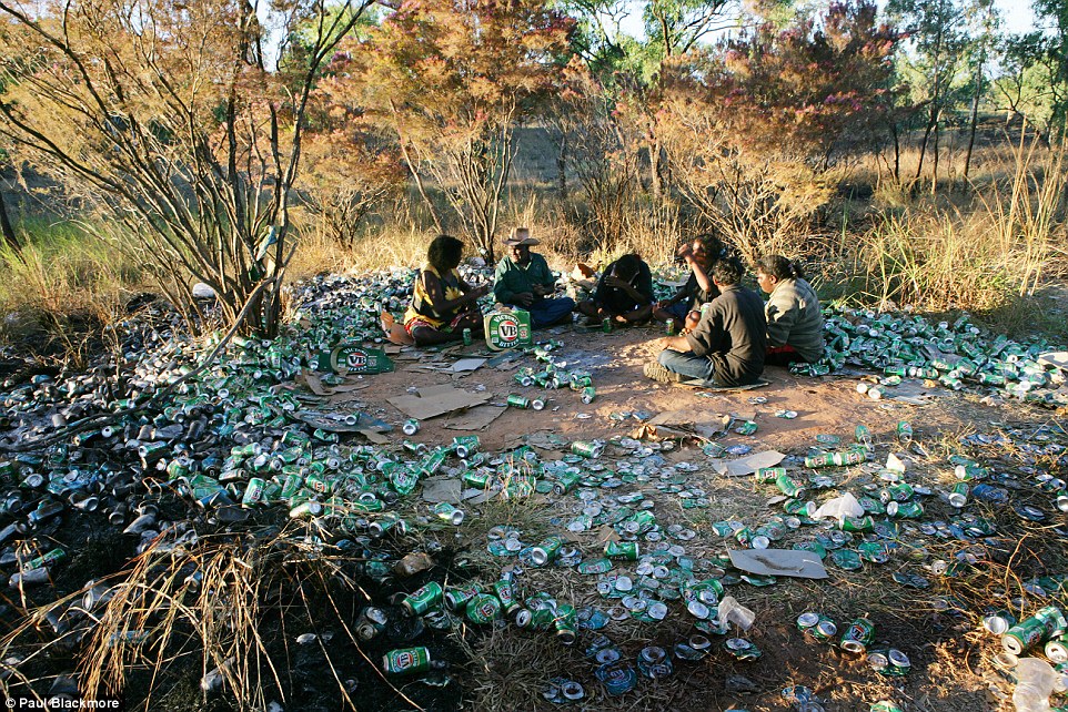 Група аборигени седят в сухи храсталаци, под тях има кръг мръсотия, осеян със стотици смачкани бирени кутии VB