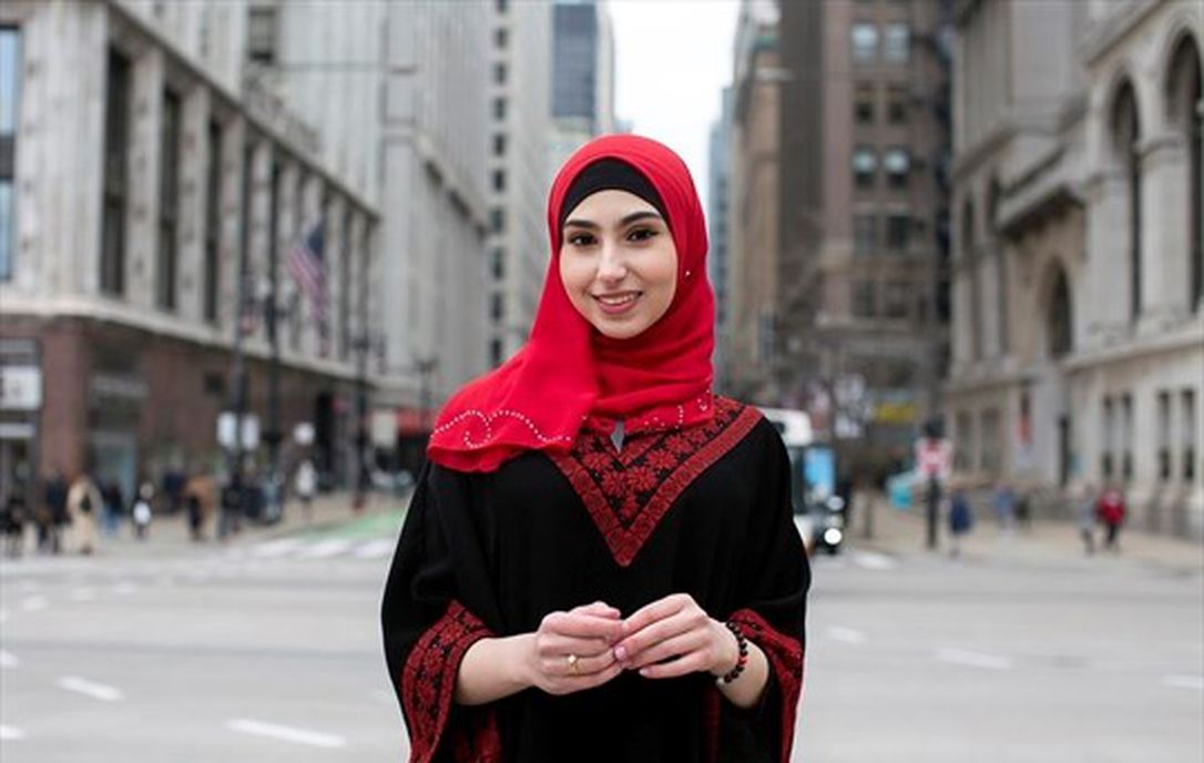 A Muslim woman wearing a red hijab, and standing on a city crosswalk with buildings behind her.