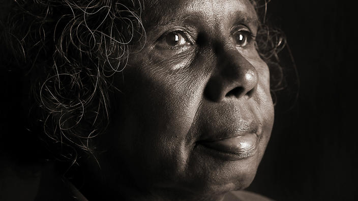 A close-up portrait of an Indigenous woman in darkness staring into the distance with a solemn expression and watery eyes