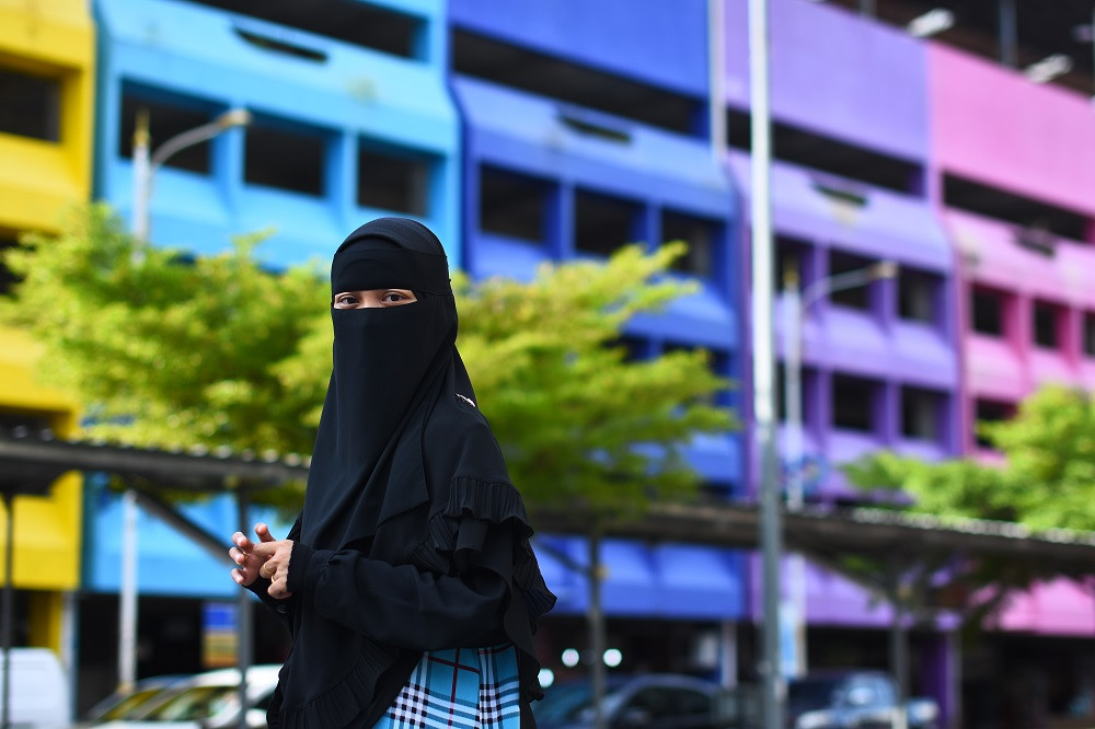 A woman wearing a black niqab stands in front of colorful buildings on the street.