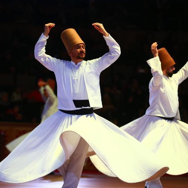 Sufi men perform their whirling dance ritual, wearing white flowing clothes and brown sikke head coverings.