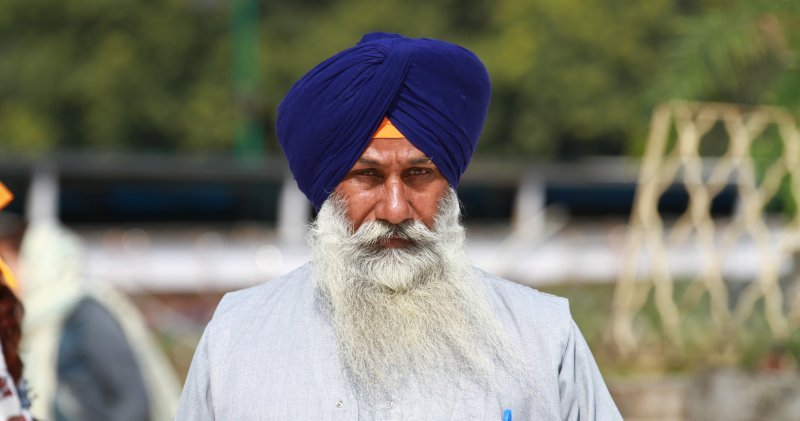 A Sikh man wearing a white top and a blue dastar turban looks into the camera.