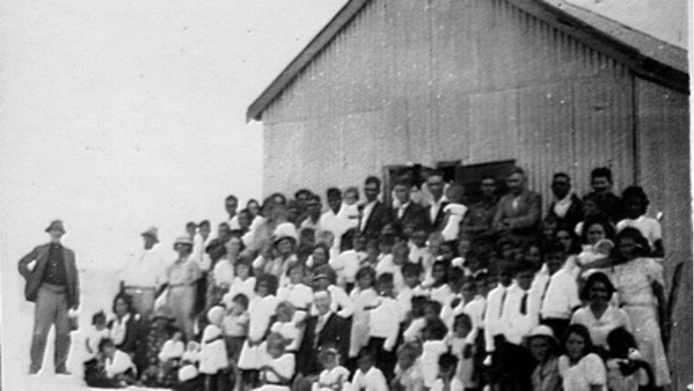 Старо черно-бяло изображение на голяма група аборигенски деца с униформи, стоящи пред сграда с бял мъж, стоящ до тях