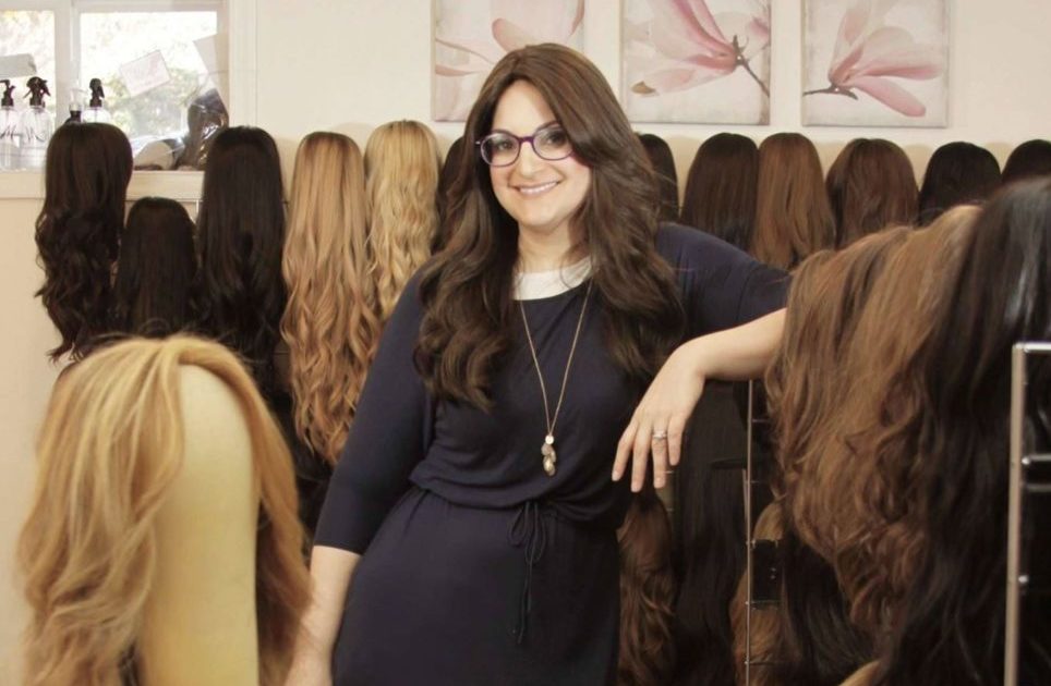 A woman with long brown hair stands in the center of a shop that has many different sheitel wigs on display.