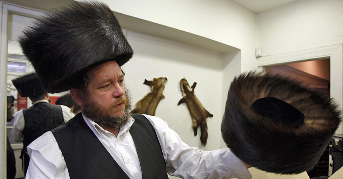 A Jewish man wearing a shtreimel holds up another sthreimel fur hat in his left hand.