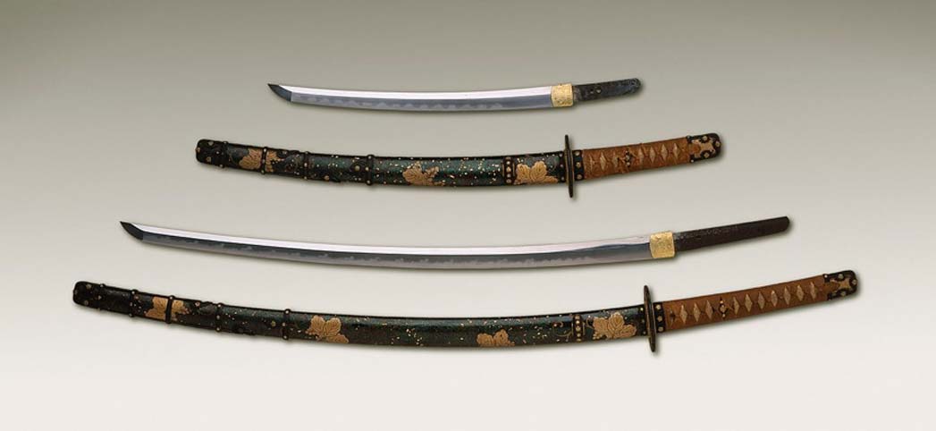 A long sword and its sheath are below a short sword and its sheath, both sheaths having intricate Japanese design, with the handles of the weapon black and gold.