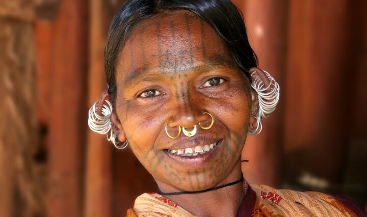 A portrait of a woman with tattooed patterns on her forehead and chin, she wears nose piercings and ear jewellery and is smiling at the camera