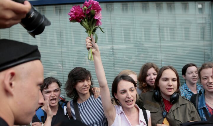 Жена, застанала сред група хора, държи с една ръка във въздуха куп розови цветя