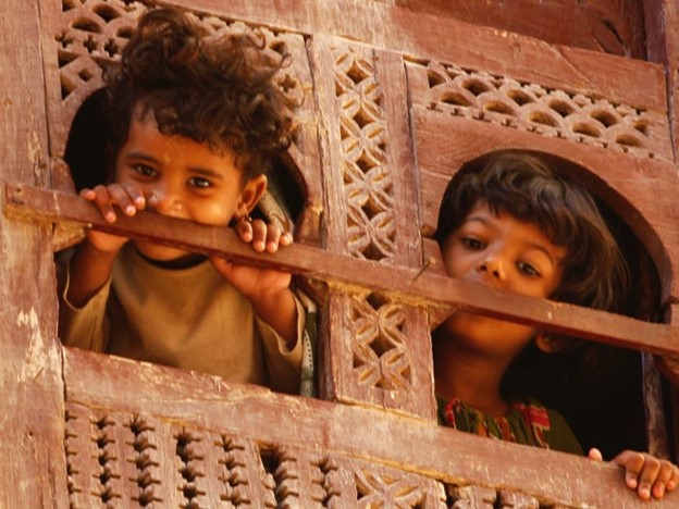 שני ילדים מתולתלים מציצים מבעד לחלון תימני מגולף מעץ לרחוב שמתחת בשיבאם.