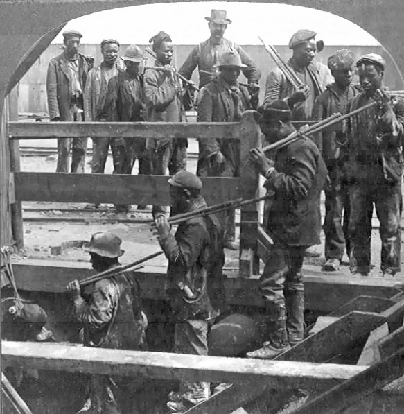 Et sort / hvidt billede af minearbejdere, der går ned ad en mineskaft af træ i 1800'erne med deres hårde hatte og pluk