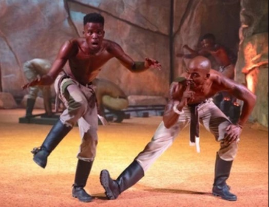 To mænd gumboot danser på scenen på en meget teatralsk måde. Den ene klapper i støvlerne, mens den anden har en fod strakt ud med fingeren på læberne