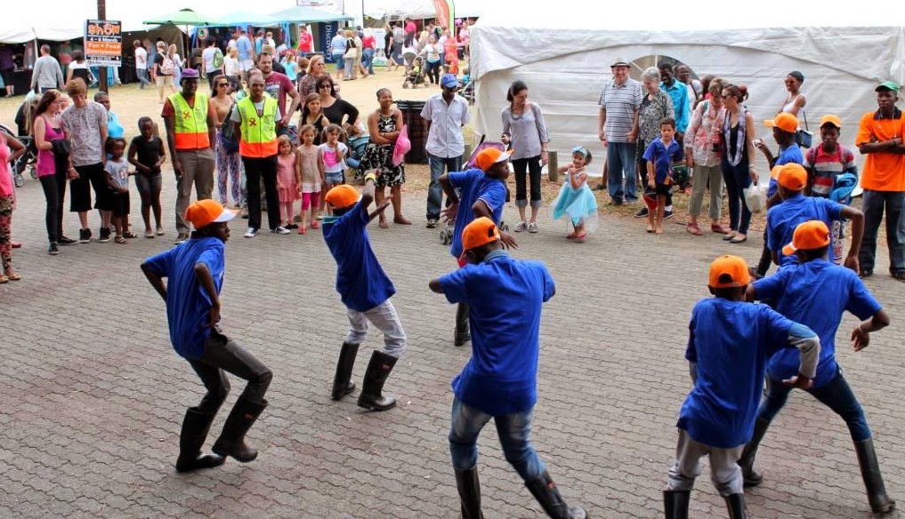 En gruppe gumboot-dansere cirkler, mens en menneskemængde ser i et ydre rum. De har blå skjorter og orange hård hatte med sorte gumboots