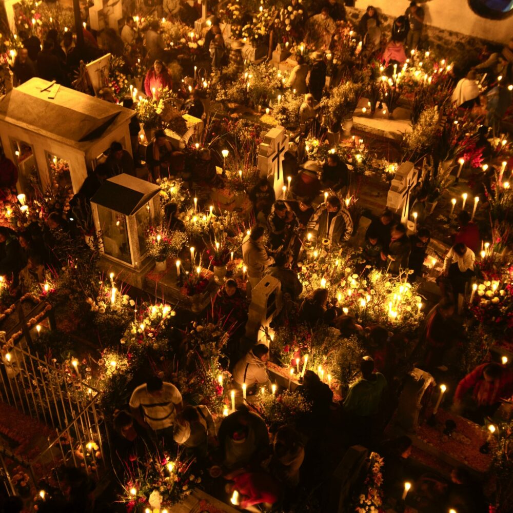 An aerial view of the grave yard in Mexico lit with candles, surrounded by flower arrangements and people praying for the souls of the departed late at night.
