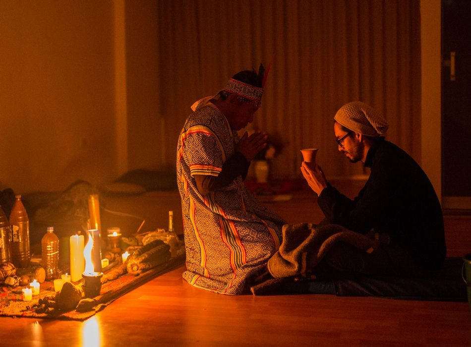 Ayahuasca Ceremony. A shaman and a participant in a black turtleneck sitting on a candlelit floor and holding a small cup with Ayahuasca.