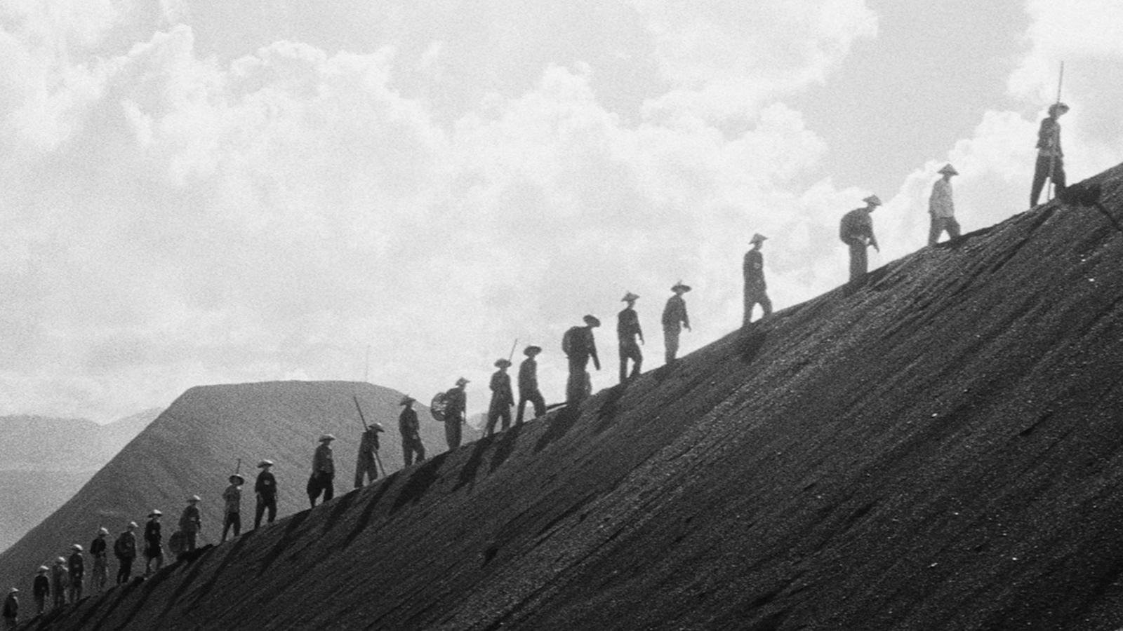 a black and white picture of men climbing a hill in a desolated place against a cloudy sky