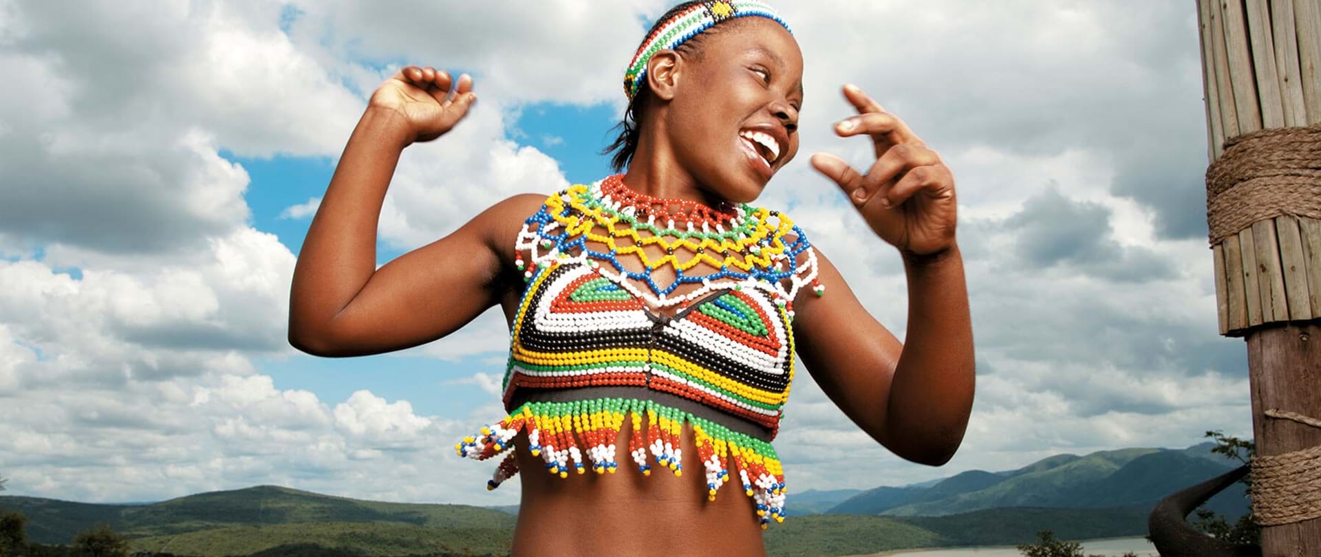 A young Zulu girl from the waist up with colourful ytraditional designs covering her torso and forehead. Her head is to the side and it looks like she is jumping against a background of a green valley and blue sky.