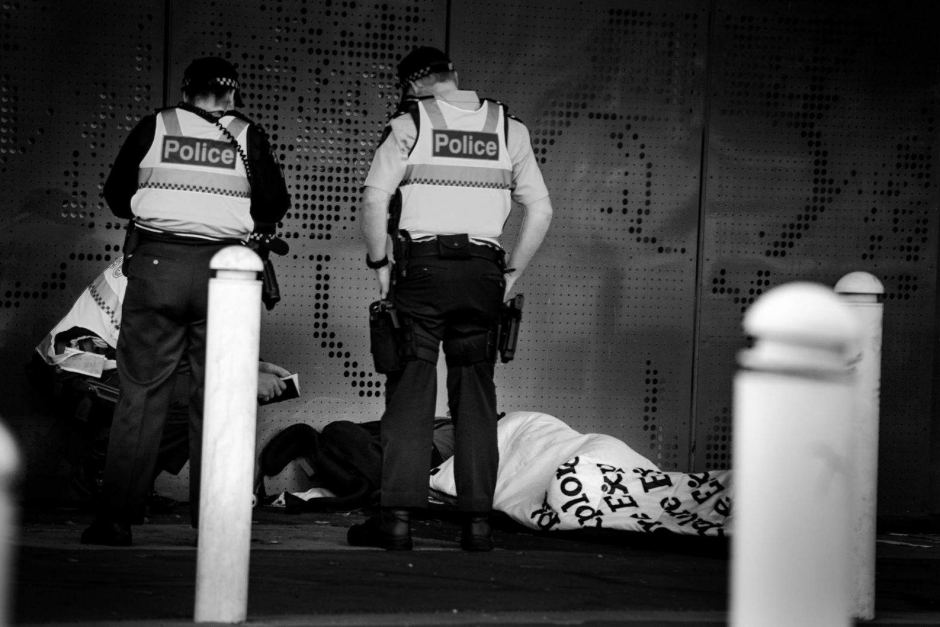 Black and white image of two police officers standing over a homeless person lying on the concrete floor in a sleeping bag