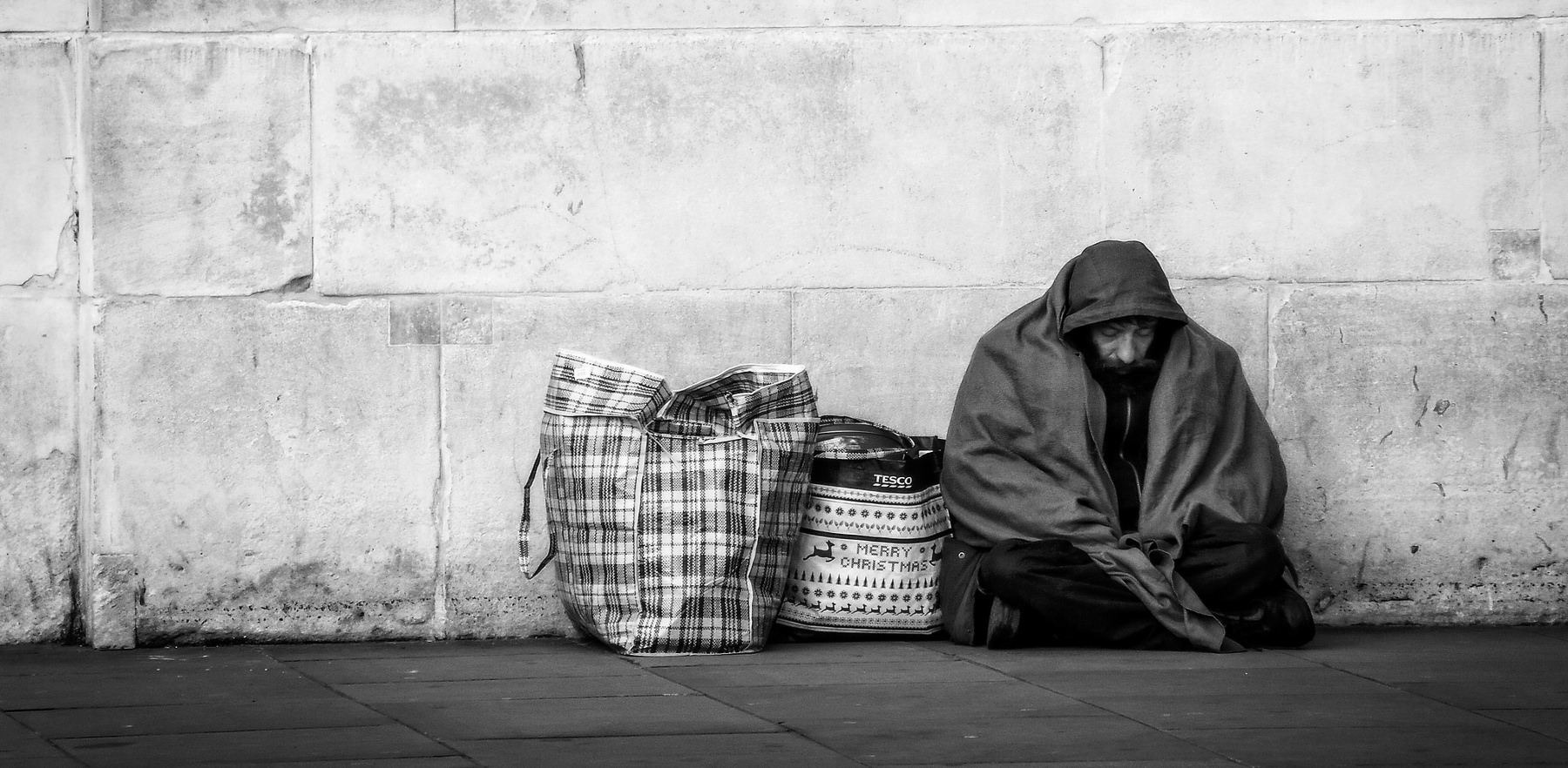 Черно -бял кадър на бездомник, прикрит в одеяло с голяма чанта до него и бетонна стена зад него