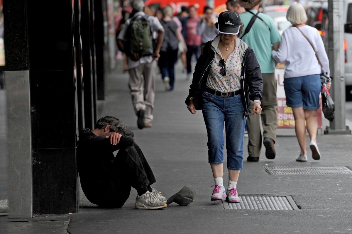 Мъж седи с глава в коленете си срещу натоварена странична разходка, докато хората минават