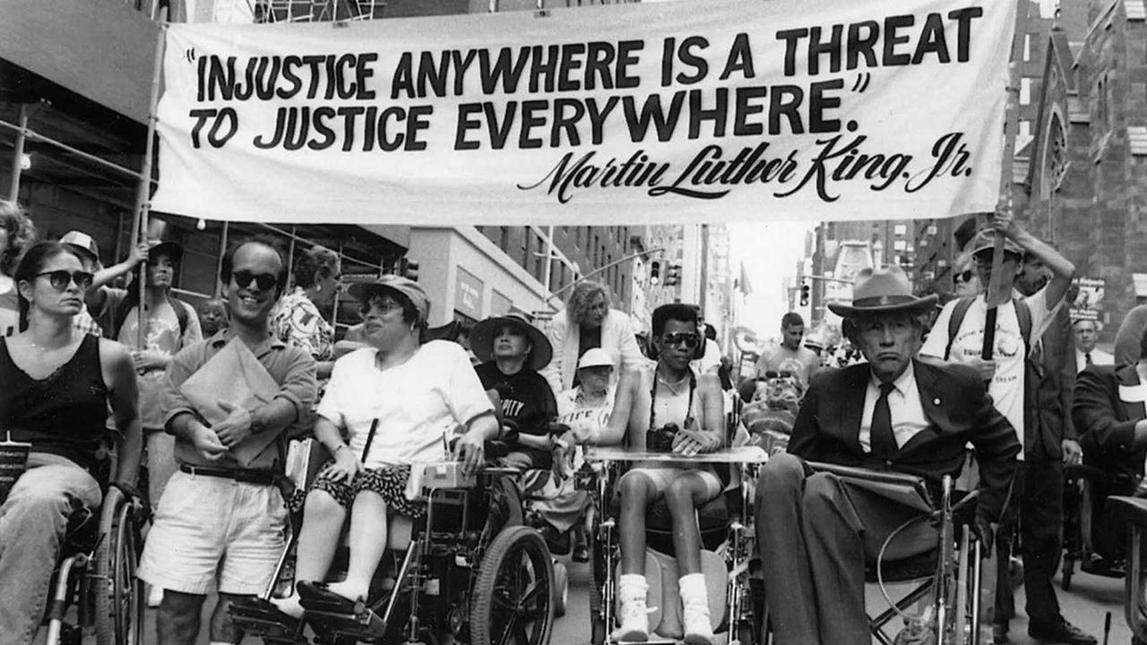 a black and white image of people with disabilities protesting with a slogan written on white sheet which says " injustice anywhere is a 