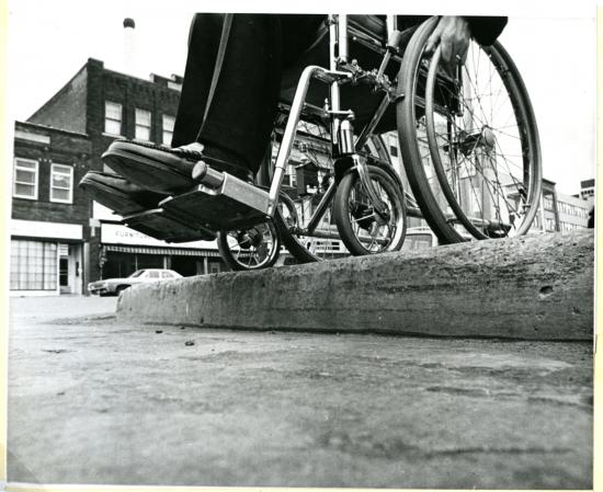 a black and white picture of a man in the wheelchair, which zooms on the wheelchair and the road that has barriers