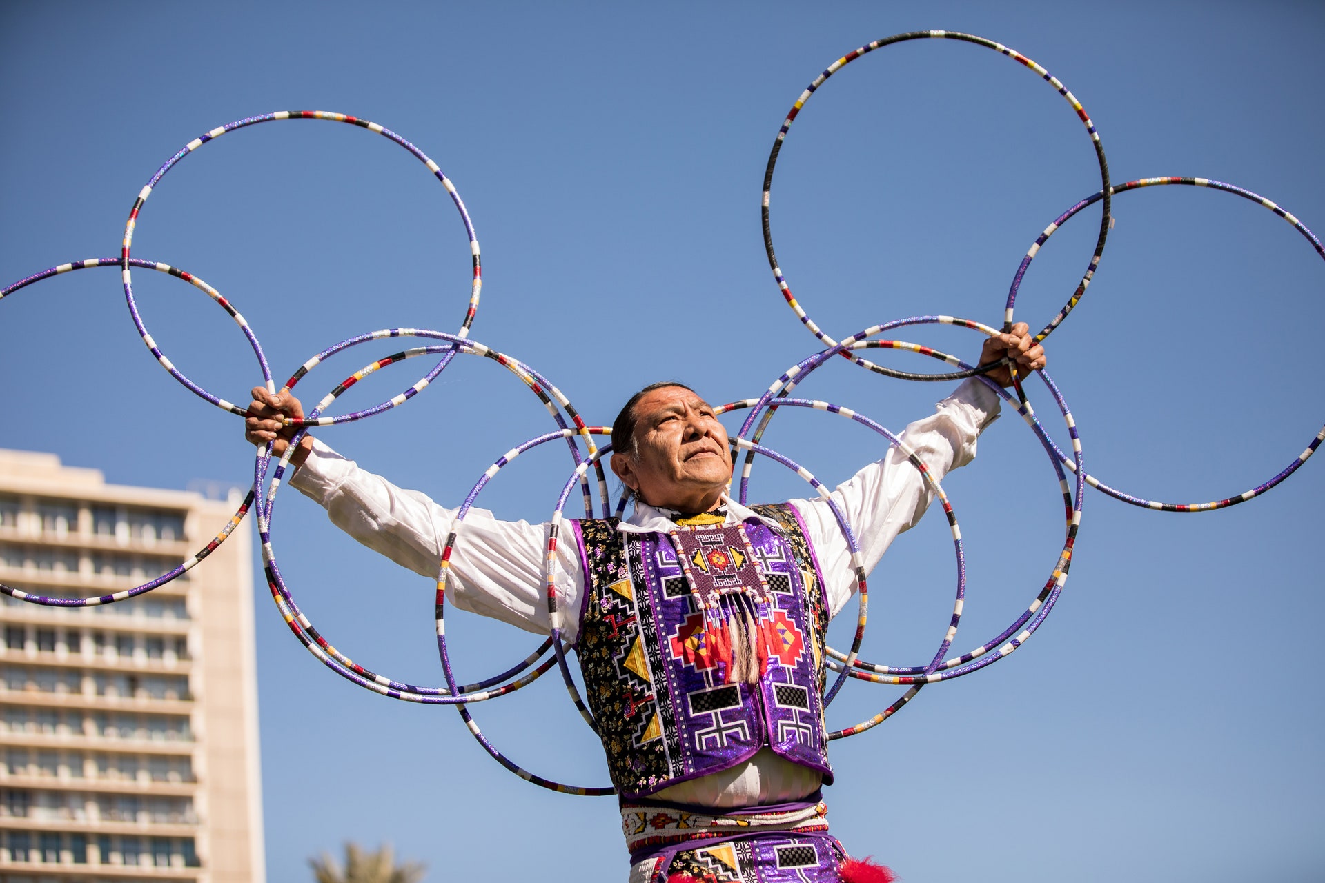 A man performing the Hoop Dance against a clear blue sky. The hoops on his arms resemble wings.