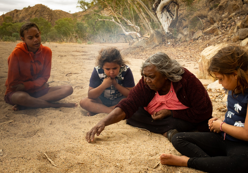 En indfødt australsk ældste kvinde sidder på sandet med tre børn og tegner billeder af Dreatime -historier i sandet med sine hænder