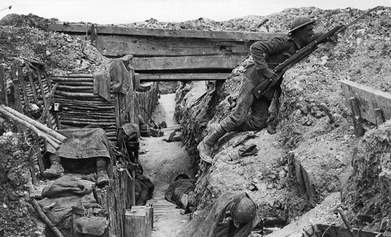 A photo of a British trench. One soldier is peeking over the edge. The trench conditions look grim and dirty
