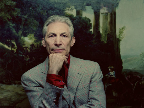 Et portræt af Charlie Watts, sandsynligvis taget i 2010'erne. Han er iført en grå jakkesæt. Et klassisk maleri er i baggrunden