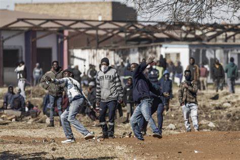 a group of African teenagers throwing stuff as part of a protest while some are standing or sitting in groups at the back o