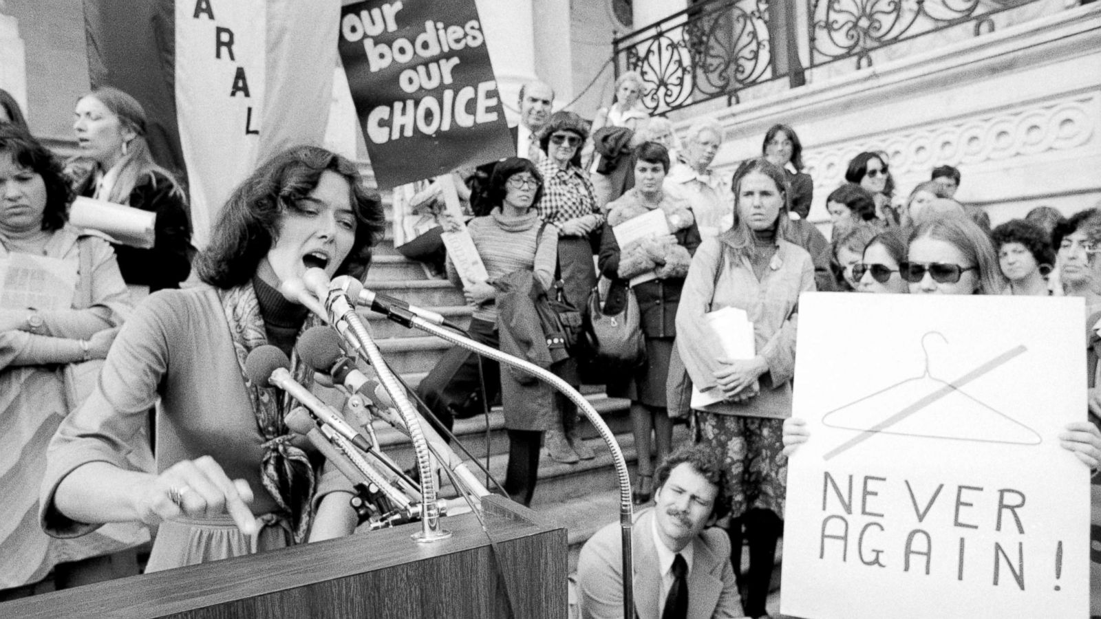 Black and white image of a courtroom in the 1970s shows a woman speaking into a microphone at the stand and people holding signs surrounding her adocating against abortion law