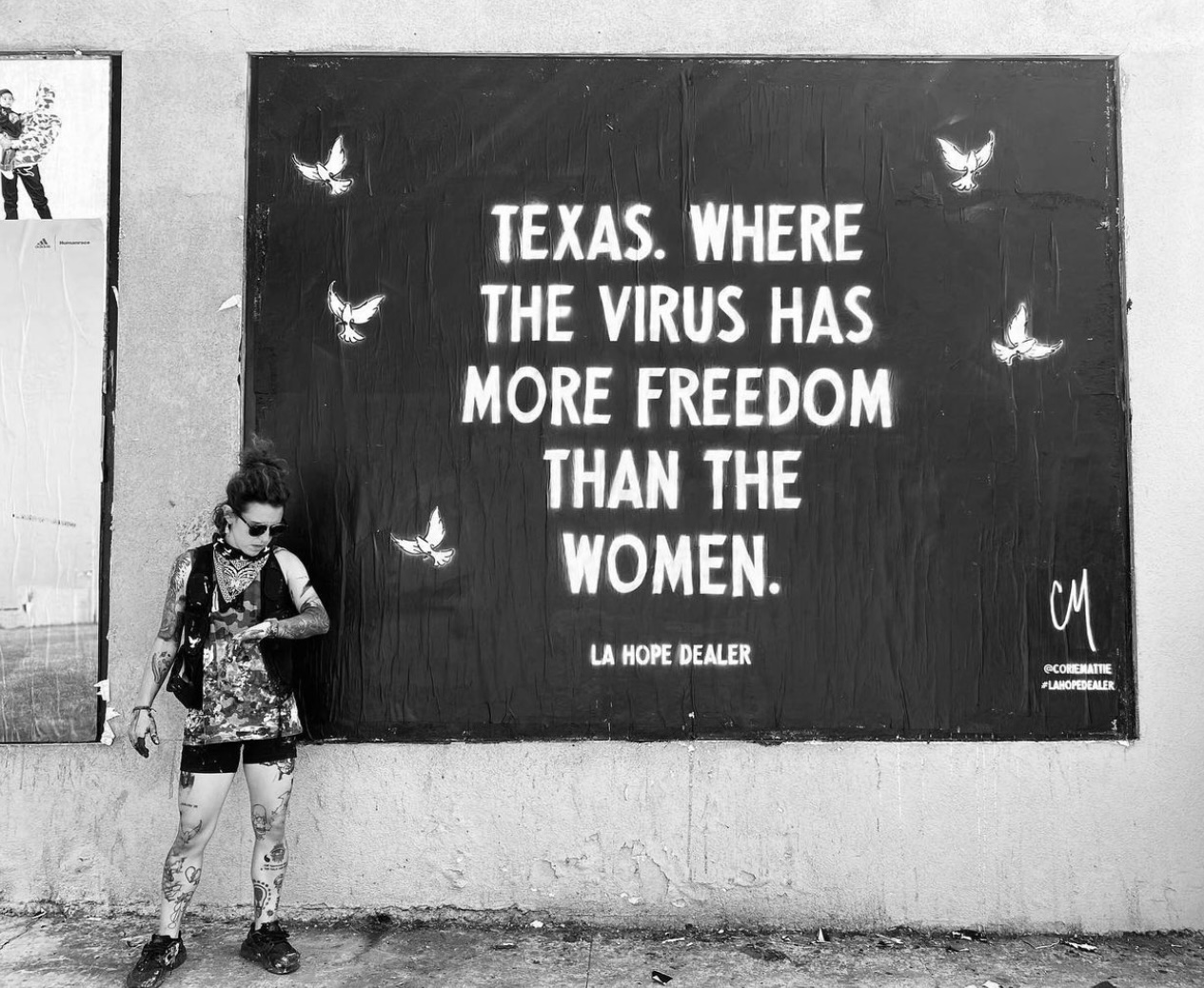 Black and white image of a woman standing next to her artwork on a street wall that says 'Texas. Where the virus has more freedom than the women'