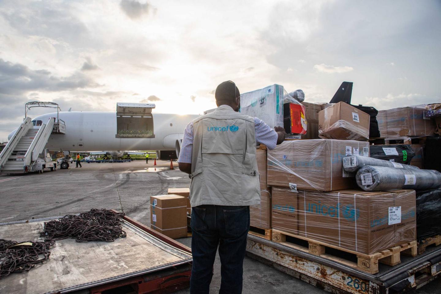 A coloured photograph pof a man standing on a landing strip with supplies provided by UNICEF for the survivors and injured of teh Haiti earthquake in 2021