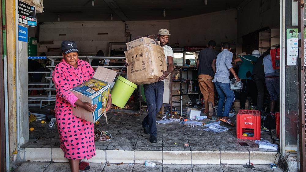 a colored image of a woman in her pink dress and a boy carrying boxes out of the  looted store as other men at the back are collecting stuff from the store 