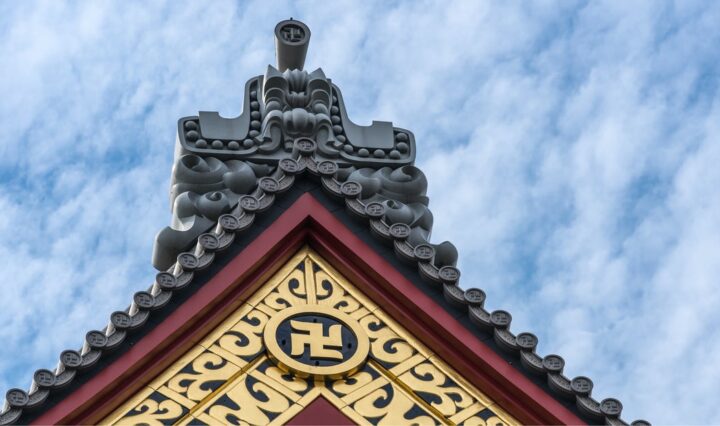 Top of a red and gold temple with a manji symbol in front of a blue clouded sky