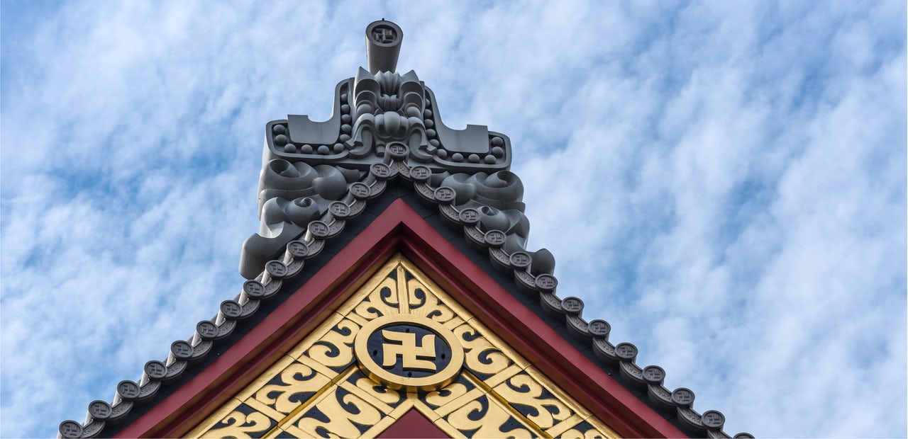 Top of a red and gold temple with a manji symbol in front of a blue clouded sky