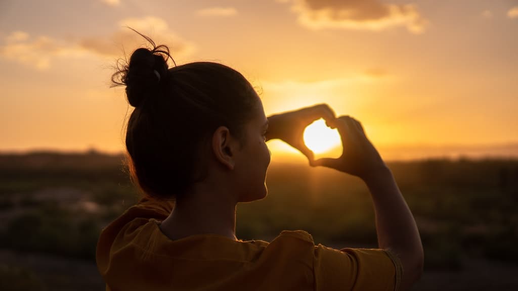 A woman forming a heart with her hands around the setting sun.