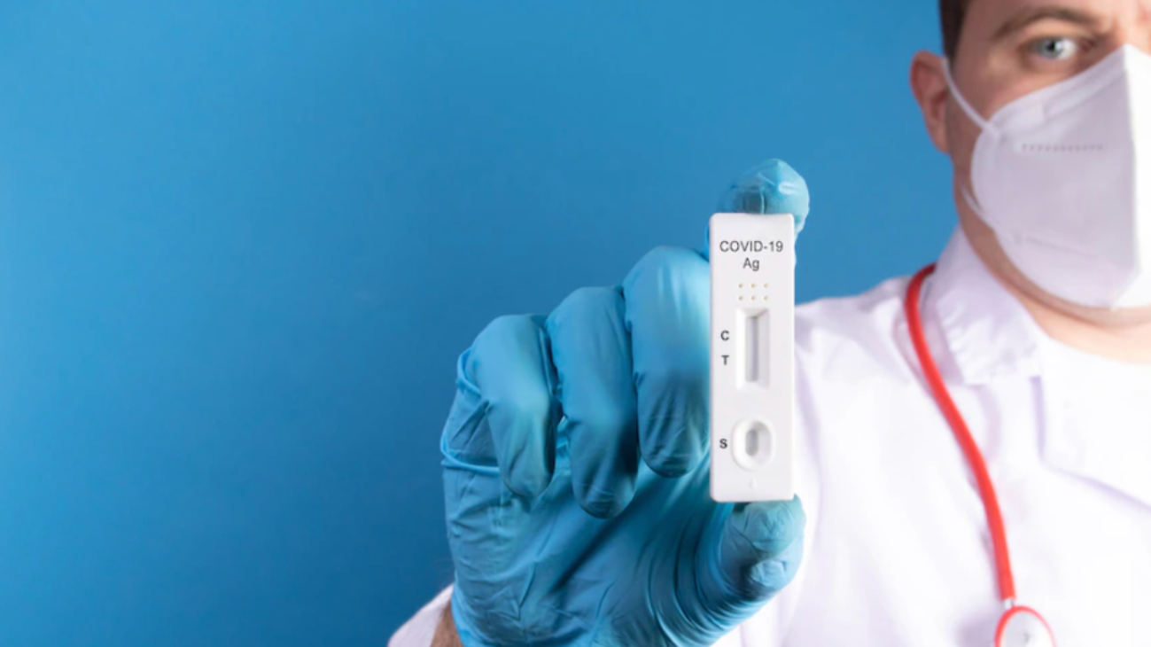 Doctor wearing a white mask and a blue glove holds a small white rapid antigen test against a blue backdrop