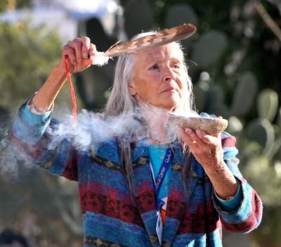Native American woman holds up an abalone shell and an eagle feather while smudge smoke from herbs rises out of the abalone shell.