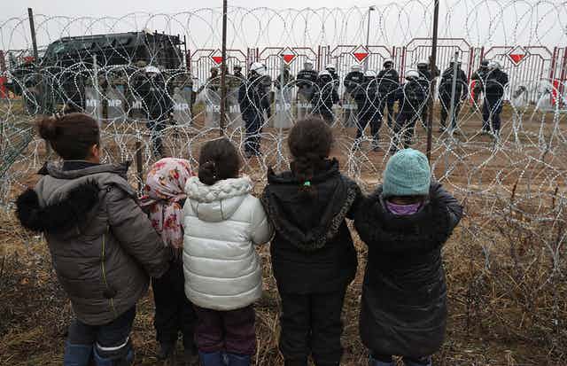 A photograph of a group of children standing at the Belarus border, staring at the Polish security forces on the other side of the barbed wire.