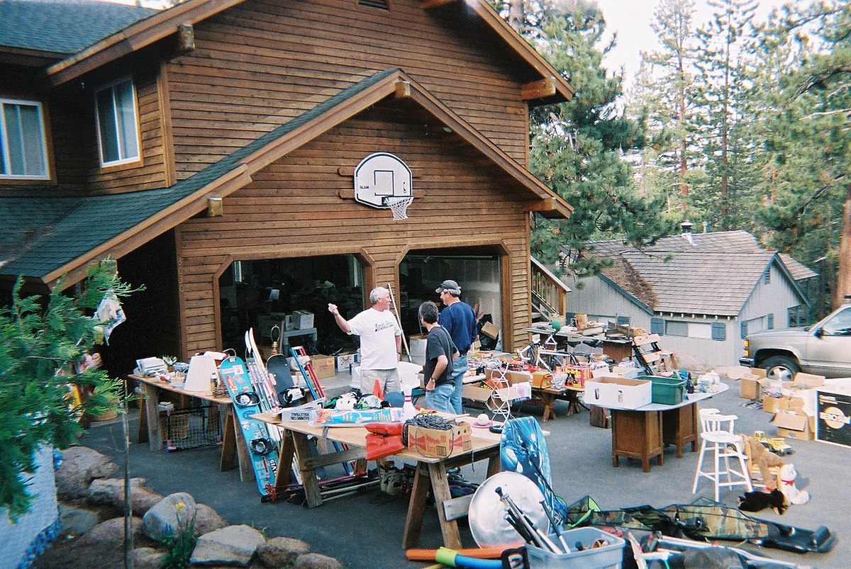 Трима мъже стоят до маси, покрити с предмети на гаражна разпродажба пред голяма дървена къща