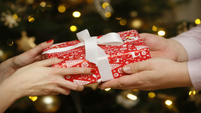 To par hænder udveksler en rød juleindpakket gave med en hvid sløjfe og juletræslys i baggrunden