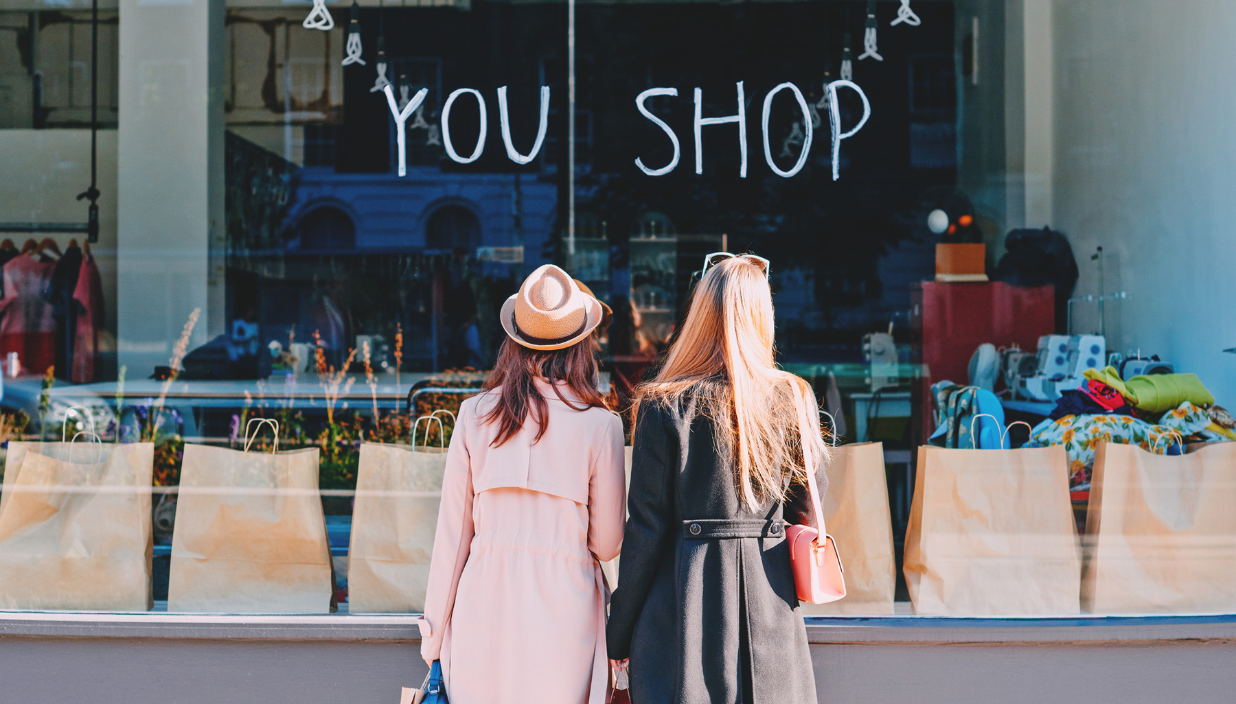 To kvinder står over for et butiksvindue foret med brune indkøbsposer og teksten 'YOU SHOP' malet på glasset