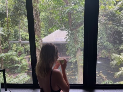 Момиче отпива от чаша чай, докато гледа през прозореца към тропическите гори Дейнтри